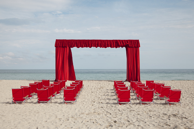 Fig. 3 Landscape with a blue sky, sea and stretch of sand where six rows of five red beach chairs are situated in front of a red theatre curtain framing the seaside view. No human figures are present.